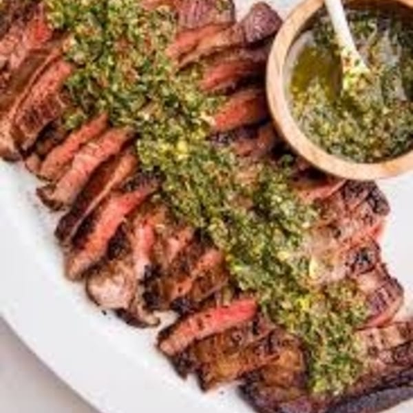 Sliced grilled steak topped with green chimichurri sauce, with a side bowl of sauce.