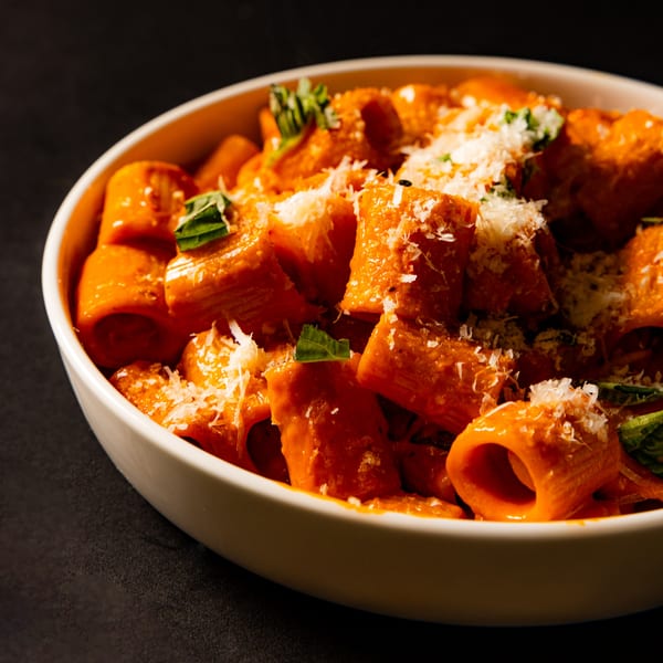 Rigatoni pasta in a creamy red sauce, topped with grated cheese and fresh basil, in a white bowl.