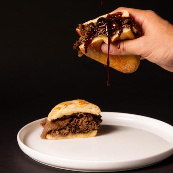 A hand holds a beef sandwich, drizzling sauce over it, with another sandwich on a white plate.