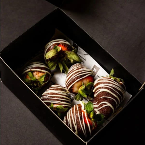 Six chocolate-covered strawberries with white drizzle and green leaves, nestled in a black box.